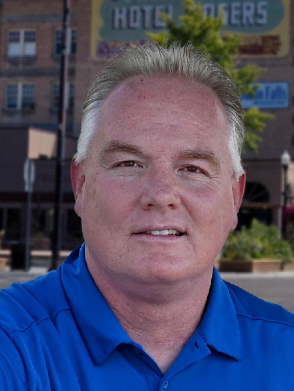 A portrait headshot photo of Brady L. Kay, a man with short, light-colored hair wearing a bright blue polo shirt smiling slightly; He is standing outdoors with a blurred city street and a brick building featuring a HOTEL sign in the background