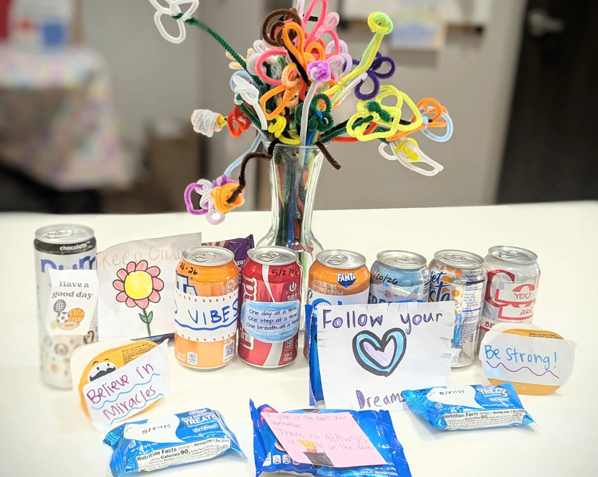 A collection of donated items on a white surface, including several cans of soda and rice cereal treats, many with handwritten encouraging notes like Believe in Miracles and Follow your dreams; In the center is a glass vase containing colorful flowers made from twisted pipe cleaners