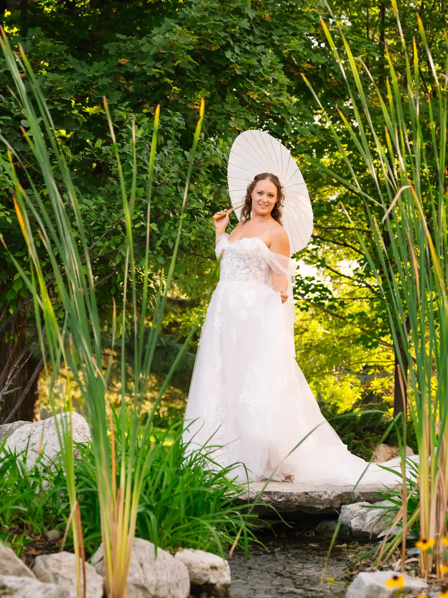 A bride in a lace off-the-shoulder wedding dress holding a white paper parasol while standing on a stone bridge in a lush outdoor garden setting.