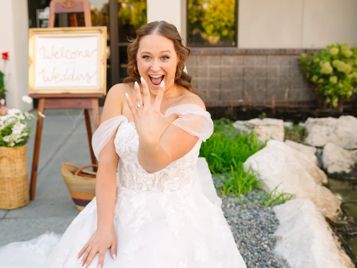 A joyful bride in a white lace wedding dress showing her diamond engagement ring and manicured nails to the camera.