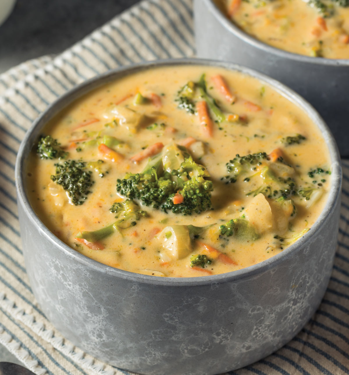 Two grey stone bowls filled with creamy, yellow-orange broccoli cheddar potato soup, topped with fresh florets and shredded carrots; The bowls rest on a blue-and-white striped linen napkin.