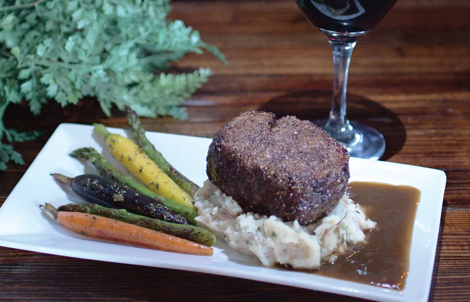 A thick, seared filet mignon rests on a bed of mashed potatoes with gravy, served alongside roasted heirloom carrots and asparagus, with a glass of red wine in the background.