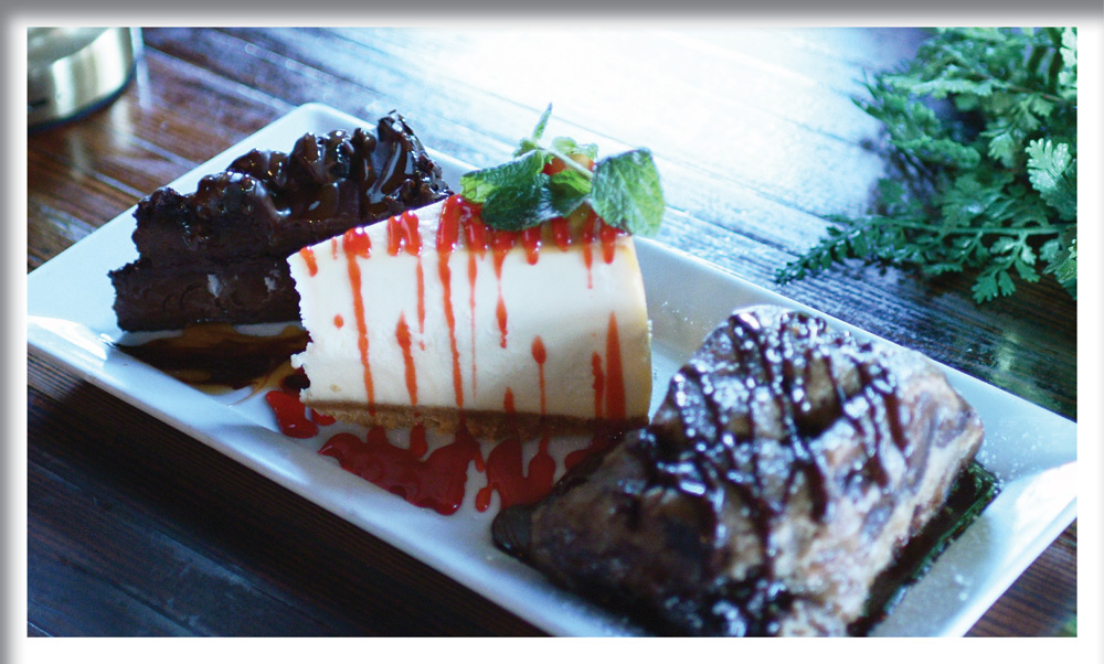 A dessert sampler plate featuring a slice of cheesecake with strawberry drizzle and mint, a rich chocolate cake, and a marbled brownie, all arranged on a rectangular white plate.