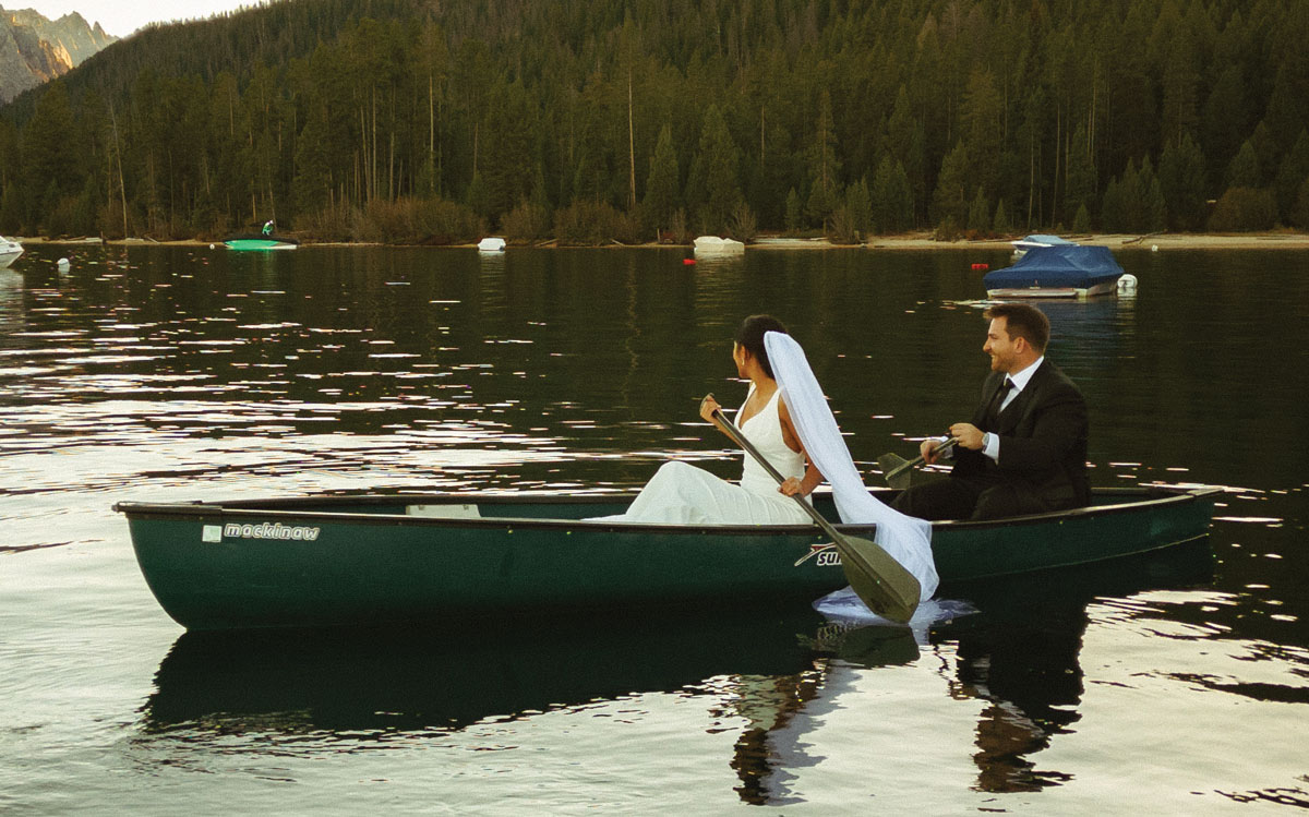 A bride and groom paddle a dark green canoe across a calm lake. Evergreen trees and a mountain peak line the shore under a soft, hazy sky. The bride sits in front, looking back.