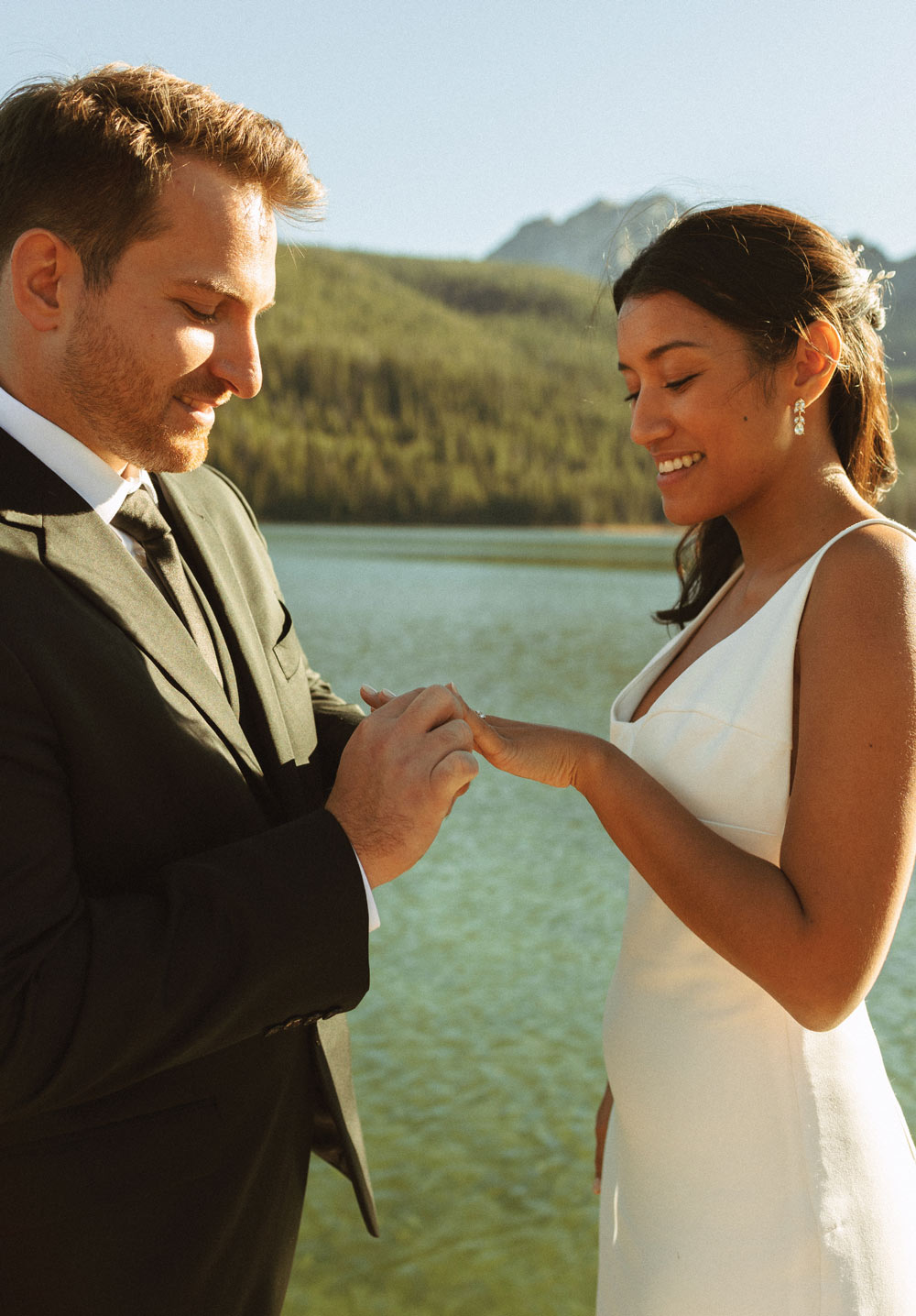 Close-up of a groom sliding a wedding ring onto the bride's finger. They are standing outdoors with a blurred blue lake and golden-lit mountains in the background.