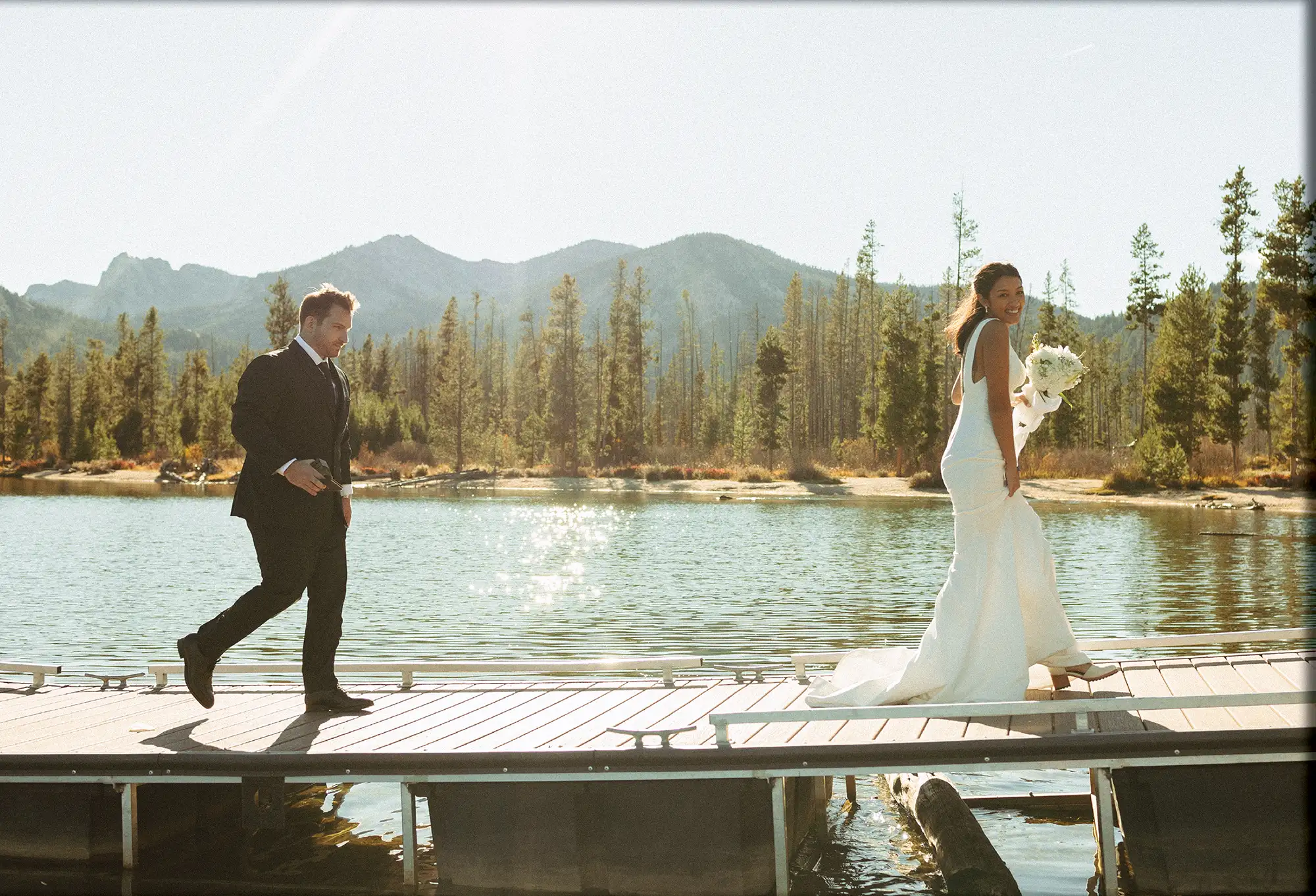 Wide shot of a bride and groom walking along a wooden pier on a sunny day. The bride carries a white bouquet and smiles at the camera, with a sparkling lake and forest behind them.