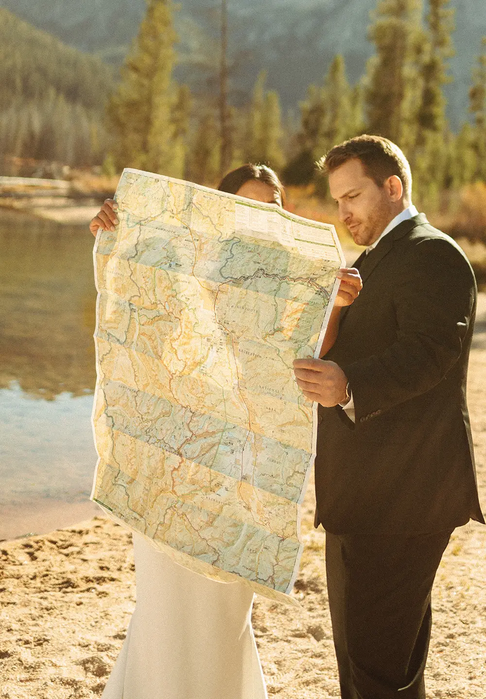 A bride and groom stand on a sandy lakeshore holding up a large paper topographical map of the Sawtooth National Forest, partially obscuring their faces in the bright sunlight.