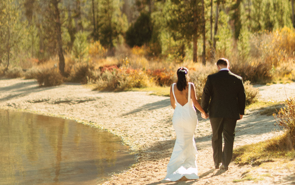 Rear view of a bride and groom walking hand-in-hand along a sandy shoreline. The bride wears a backless white gown, and the scene is surrounded by autumn-colored brush and pine trees.