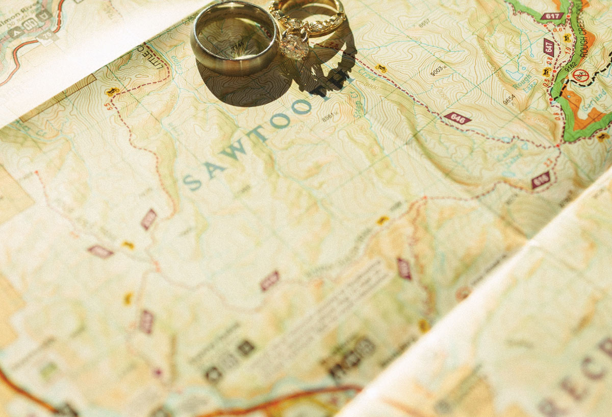 Close-up of two gold wedding bands resting on a folded topographical map. The word "SAWTOOTH" is clearly visible on the map beneath the rings, which cast soft shadows in the sun.