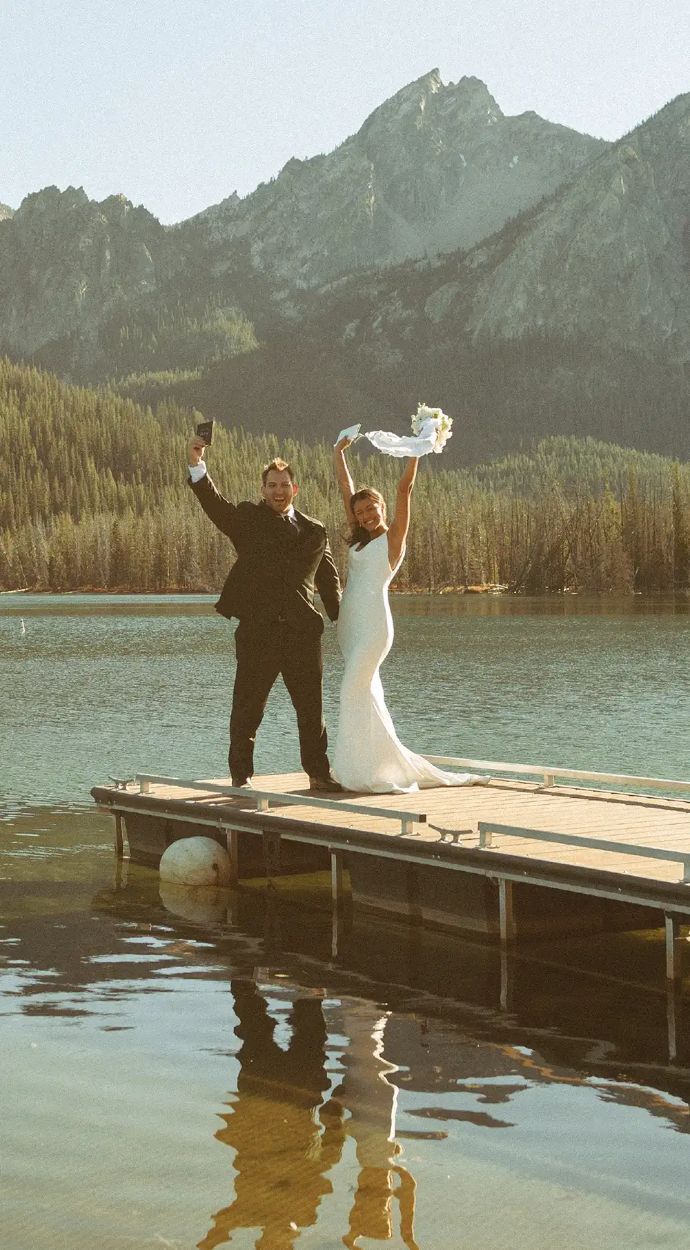 A bride and groom stand at the end of a wooden dock, raising their arms in celebration. Jagged, rocky mountain peaks rise sharply behind them over the still water of the lake.
