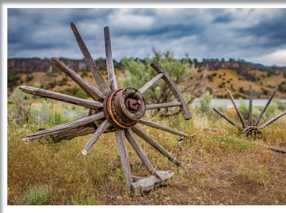 Weathered wooden wagon wheels with missing spokes lie abandoned in a grassy field. In the distance, a river flows beneath a line of rocky bluffs under a cloudy, overcast sky.