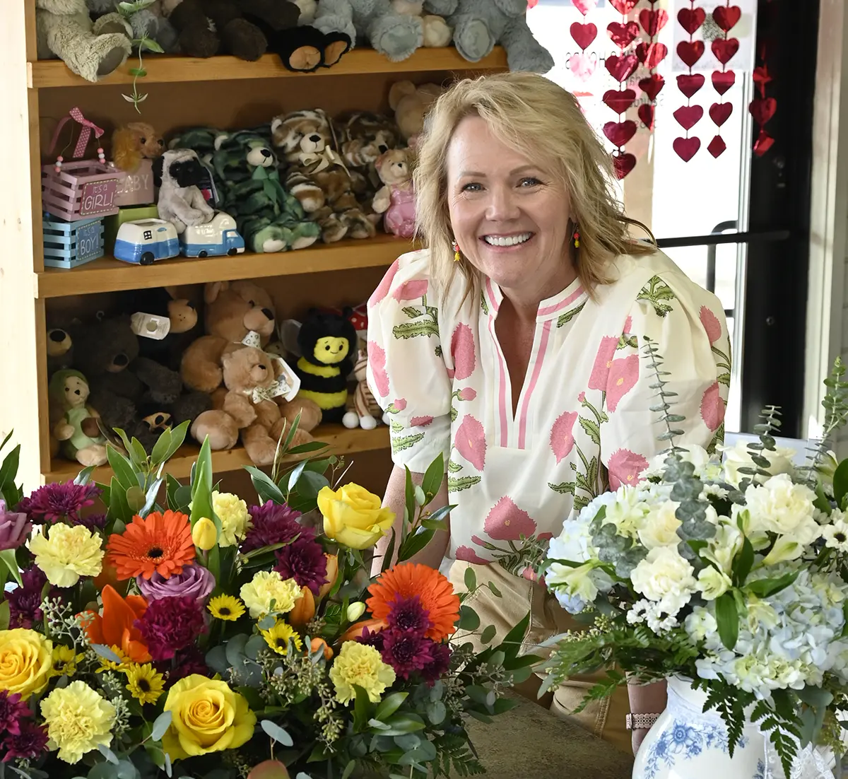 Betsy Stevens standing behind a counter with vibrant floral arrangements, featuring roses, gerbera daisies, and hydrangeas, in a gift shop filled with plush teddy bears