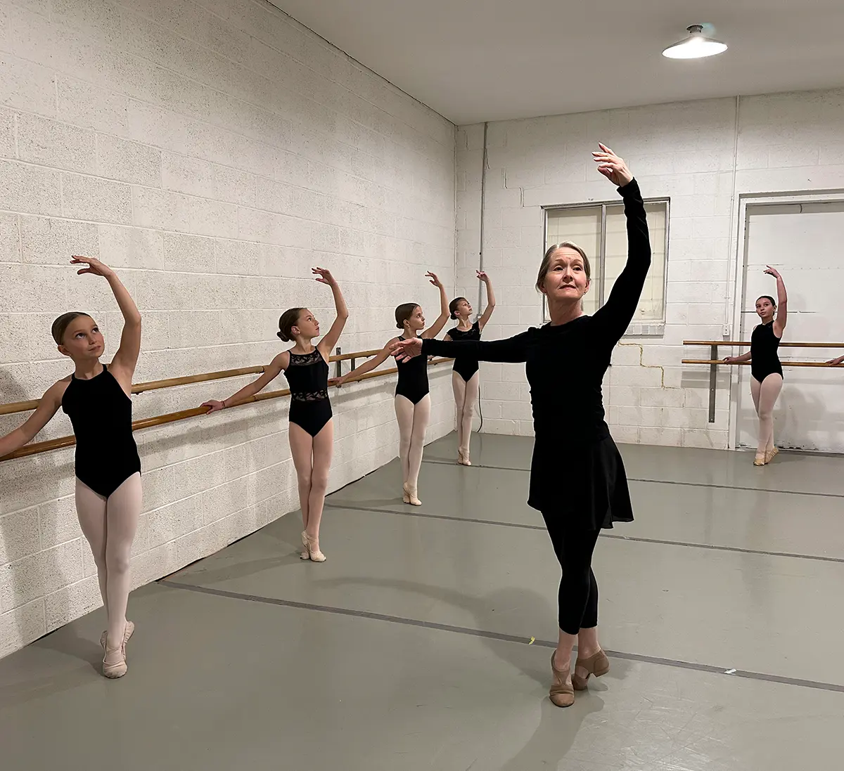 A professional ballet instructor in black dancewear leading a class of five young students in black leotards and pink tights at a wooden barre in a minimalist dance studio