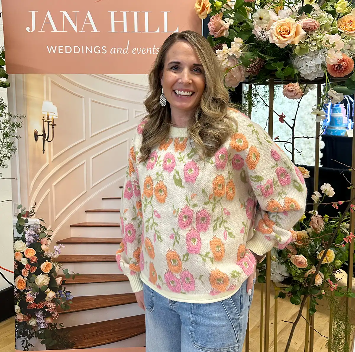 Jana Hill posing in front of a floral-decorated event backdrop featuring a grand staircase and peach-colored roses