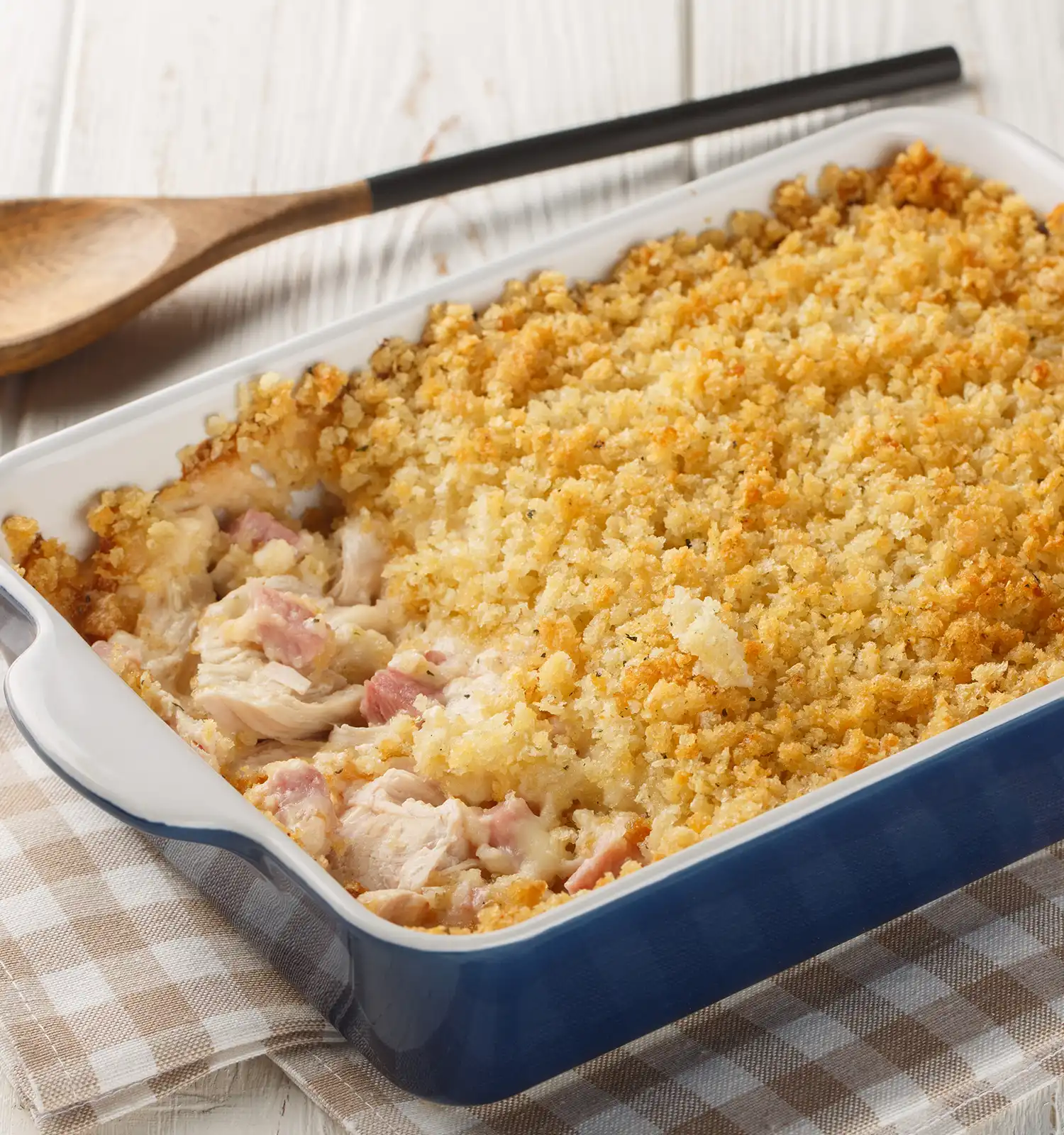 Chicken Cordon Bleu casserole in a blue baking dish with a golden-brown breadcrumb topping. A portion is removed to show chunks of chicken and ham in a creamy sauce. The dish sits on a checkered napkin next to a wooden spoon.