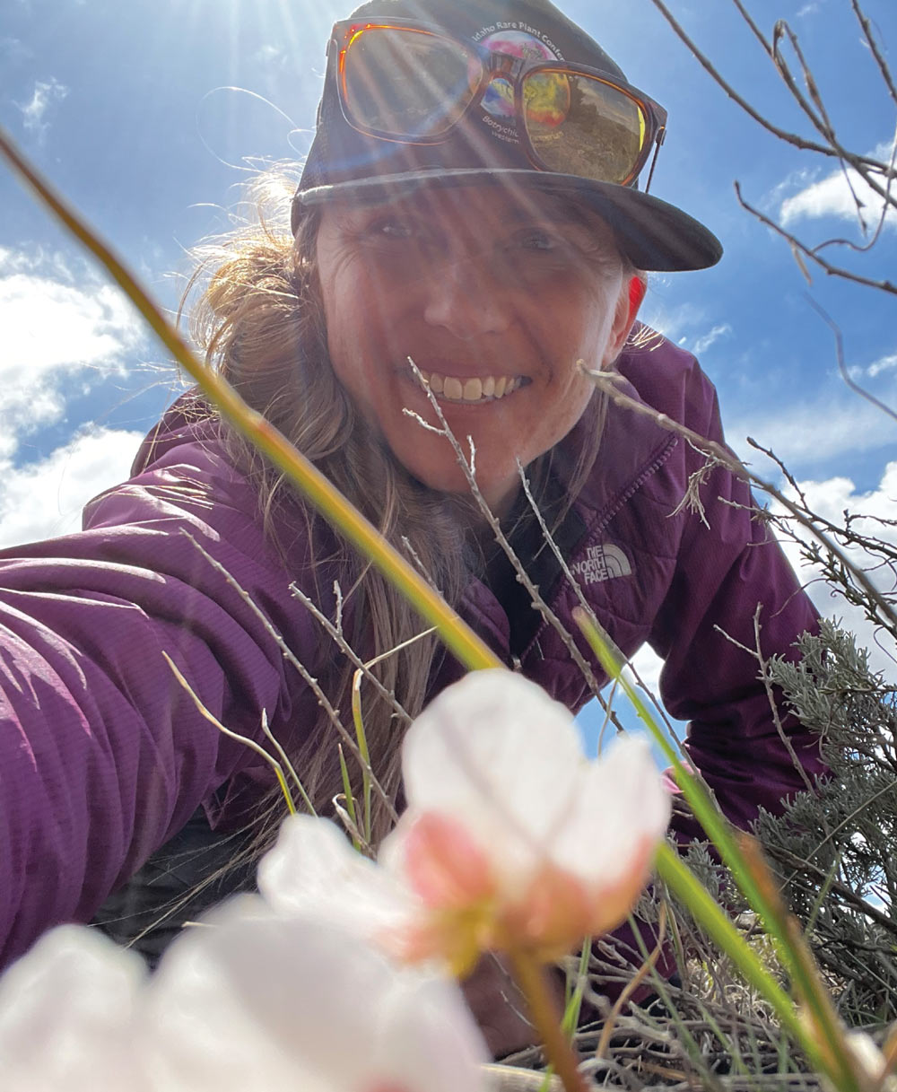 A smiling woman in a purple jacket and "Idaho Rare Plant Conference" hat leans down toward the camera. Out-of-focus white flowers are in the immediate foreground under a bright sun.