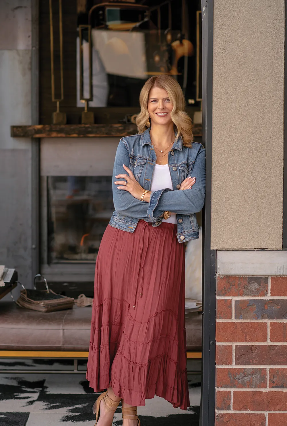 Lisa Spray in a denim jacket and tiered maroon maxi skirt leans against a doorway. She has her arms crossed in a stylish, modern interior with brick accents.