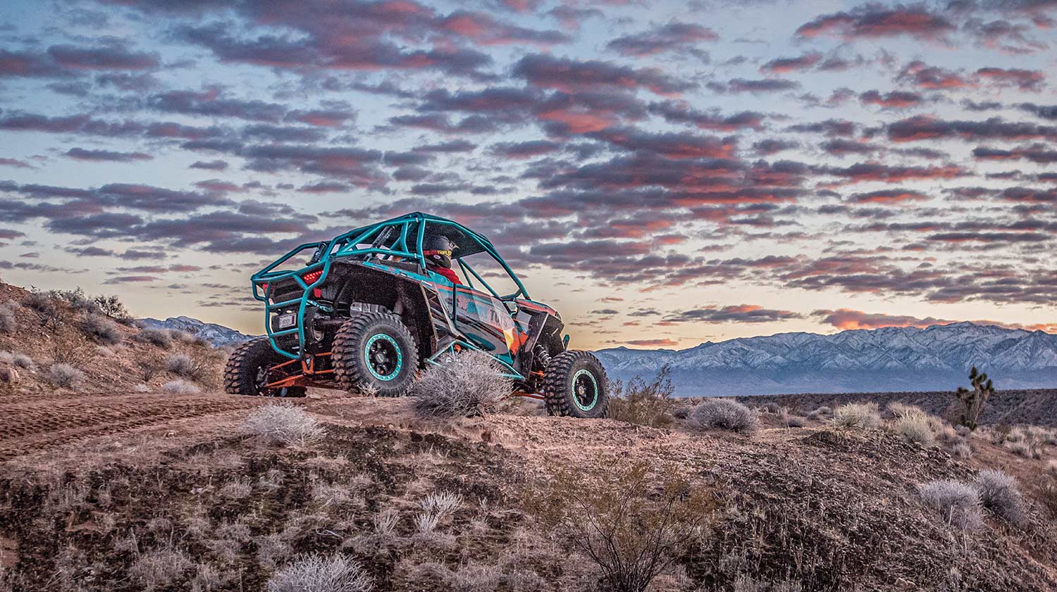 A blue off-road vehicle sits on a desert ridge against a sunset sky.