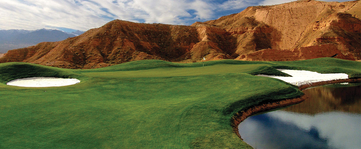 Wide view of a golf green with a sand trap and pond beneath red hills.