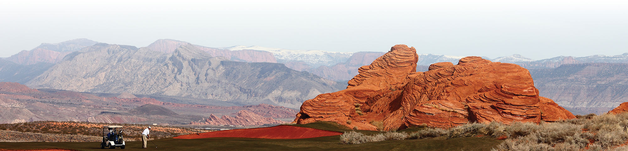Panoramic view of a golfer near layered red rock formations and distant snowy mountains.