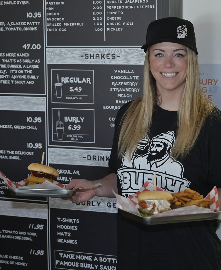 Hayley Smith smiles while holding two trays of burgers in front of a menu.