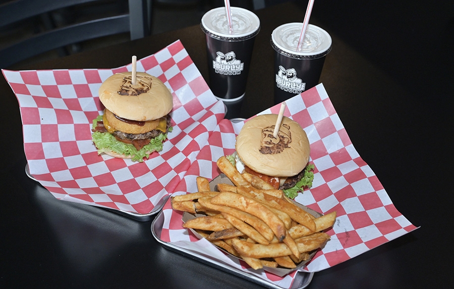 Two gourmet burgers and a side of fries on checkered paper with two drinks.