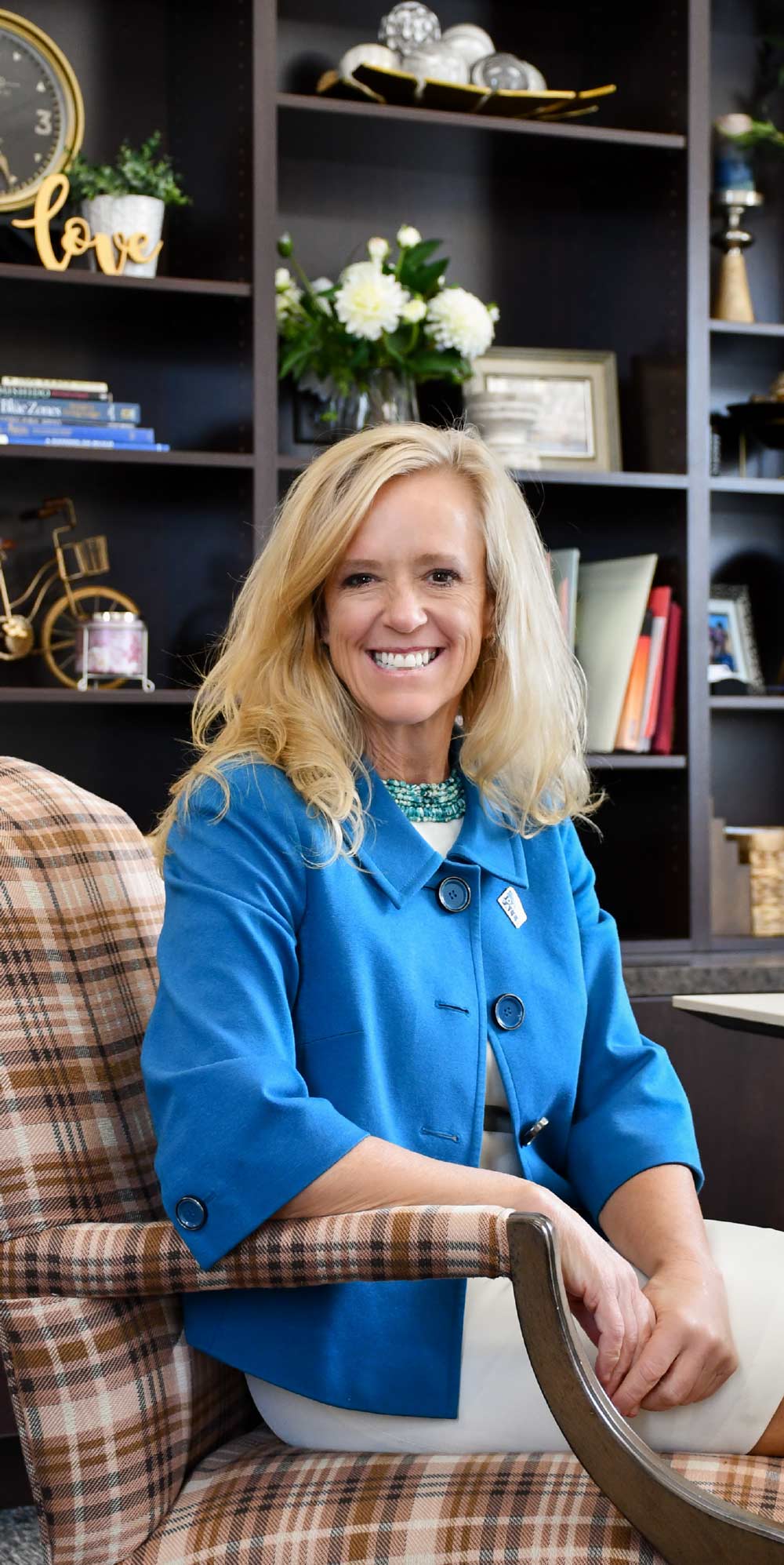 A smiling blonde woman in a bright blue jacket and turquoise necklace sits in a plaid armchair in front of a dark bookshelf with decor.