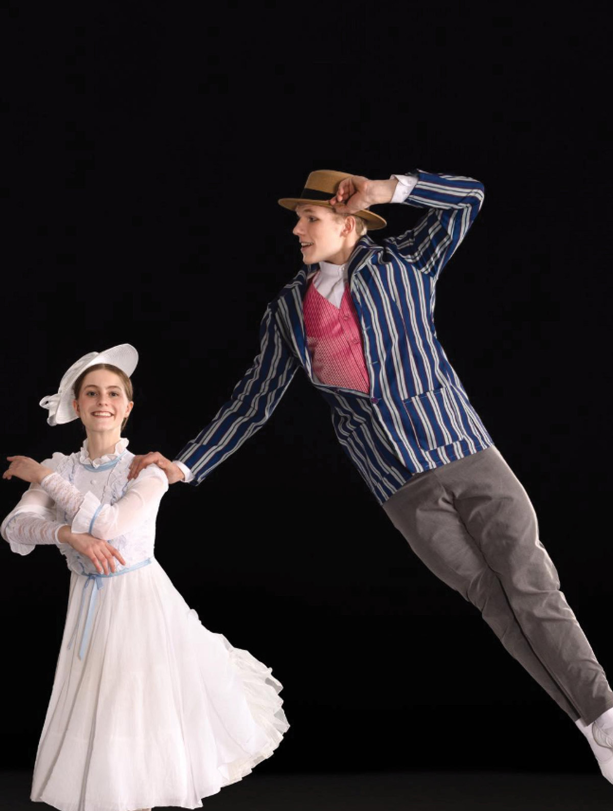 A female dancer in a white Mary Poppins-style dress attire while a male dancer is mid-air in a horizontal leap touching the female dancer's shoulder