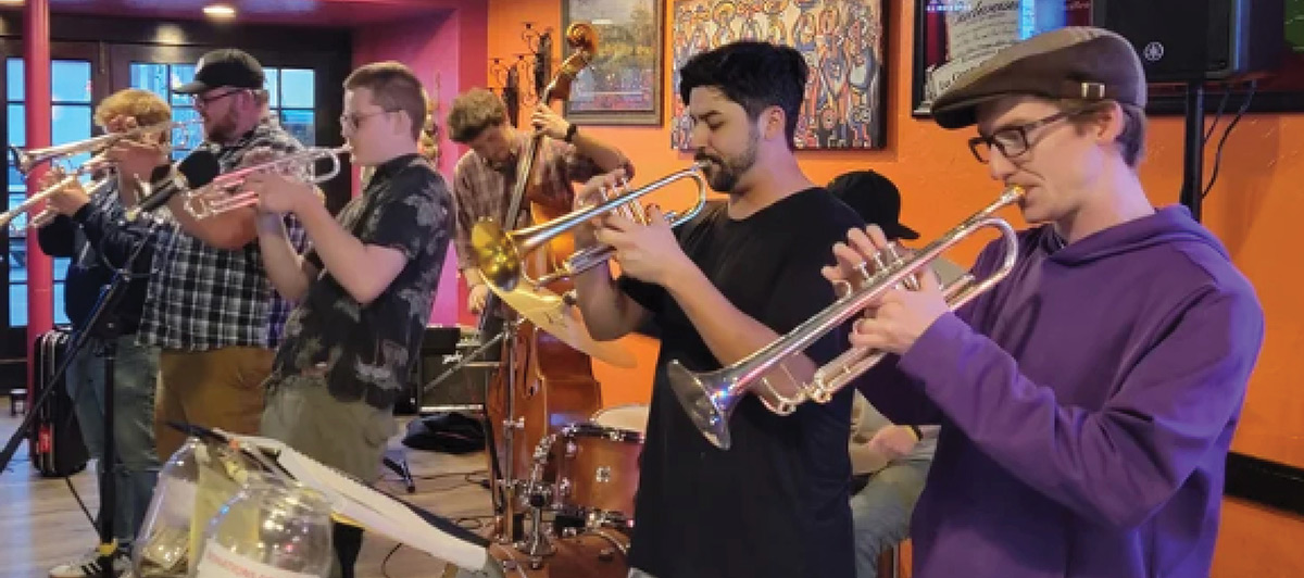 A wide, indoor shot of a jazz ensemble performing in a dimly lit venue with orange and pink walls; Each individual is focused on playing their instruments respectively
