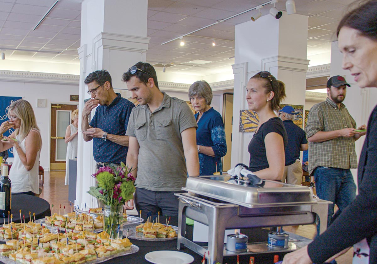 People at an indoor reception gather around a buffet table with trays of appetizers and a chafing dish. The setting is a bright, white-pillared room with artwork on the walls and attendees socializing.