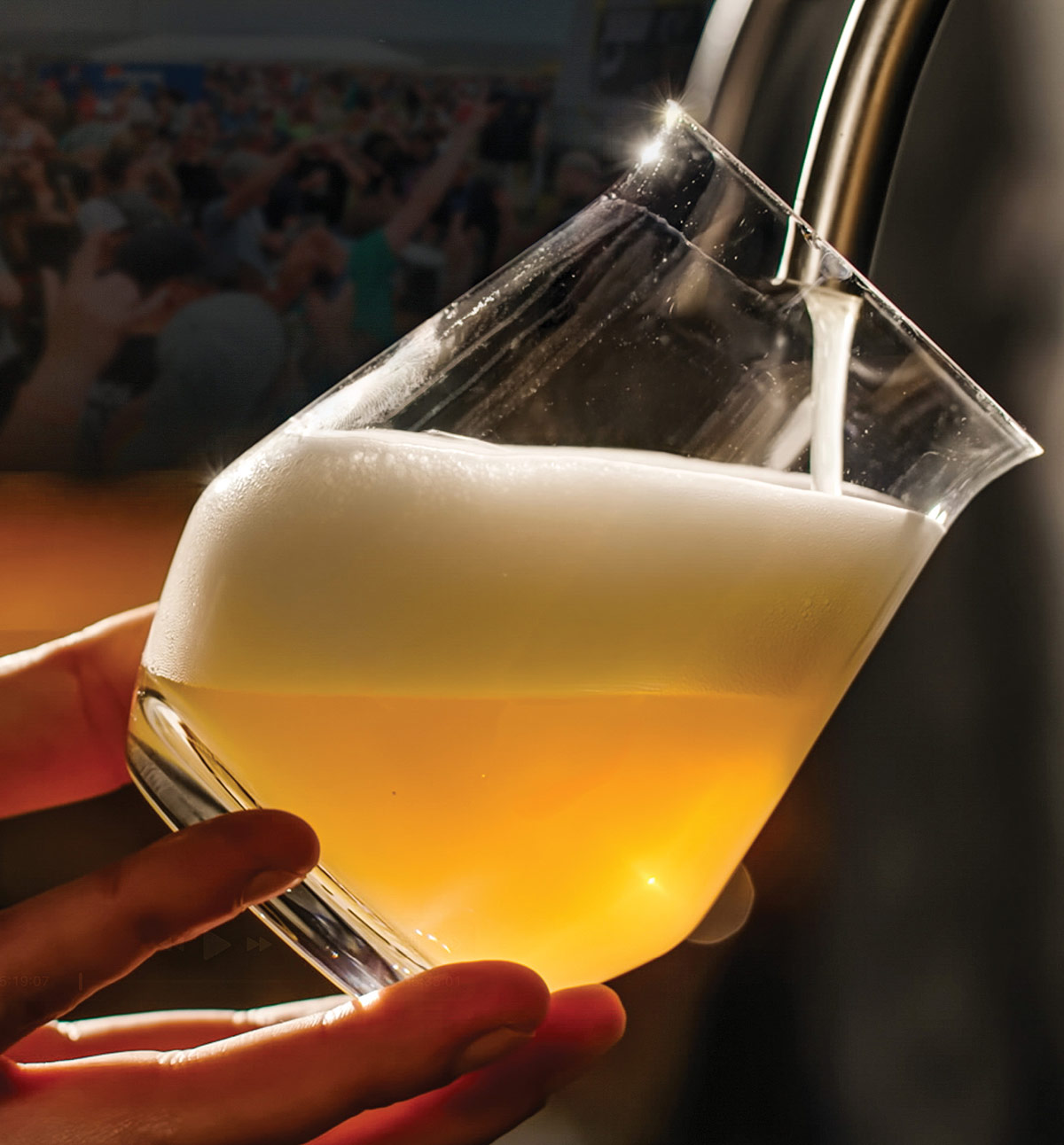Close-up of golden beer being poured from a tap into a tilted glass, creating a thick white frothy head. A hand holds the glass under dramatic lighting against a blurred background.