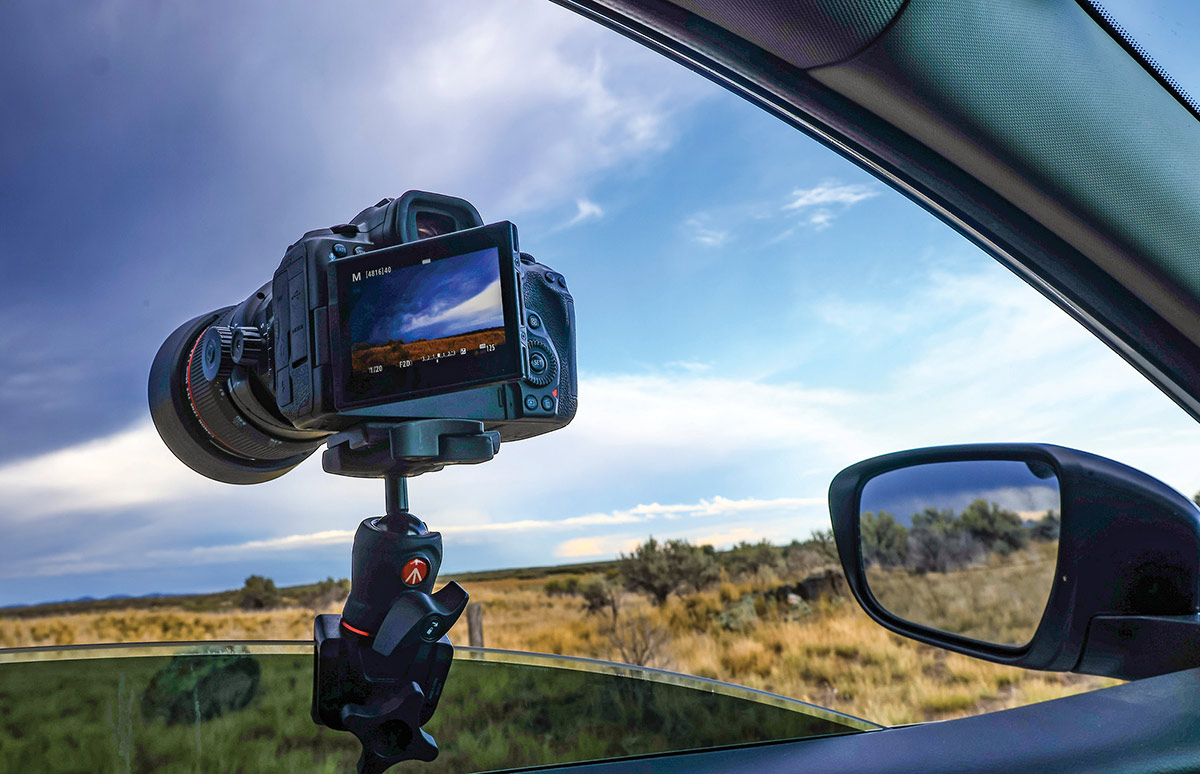 Camera mounted on a car window with a tripod head, capturing a landscape of fields under dramatic clouds. The camera's LCD screen displays the live view of the scene.