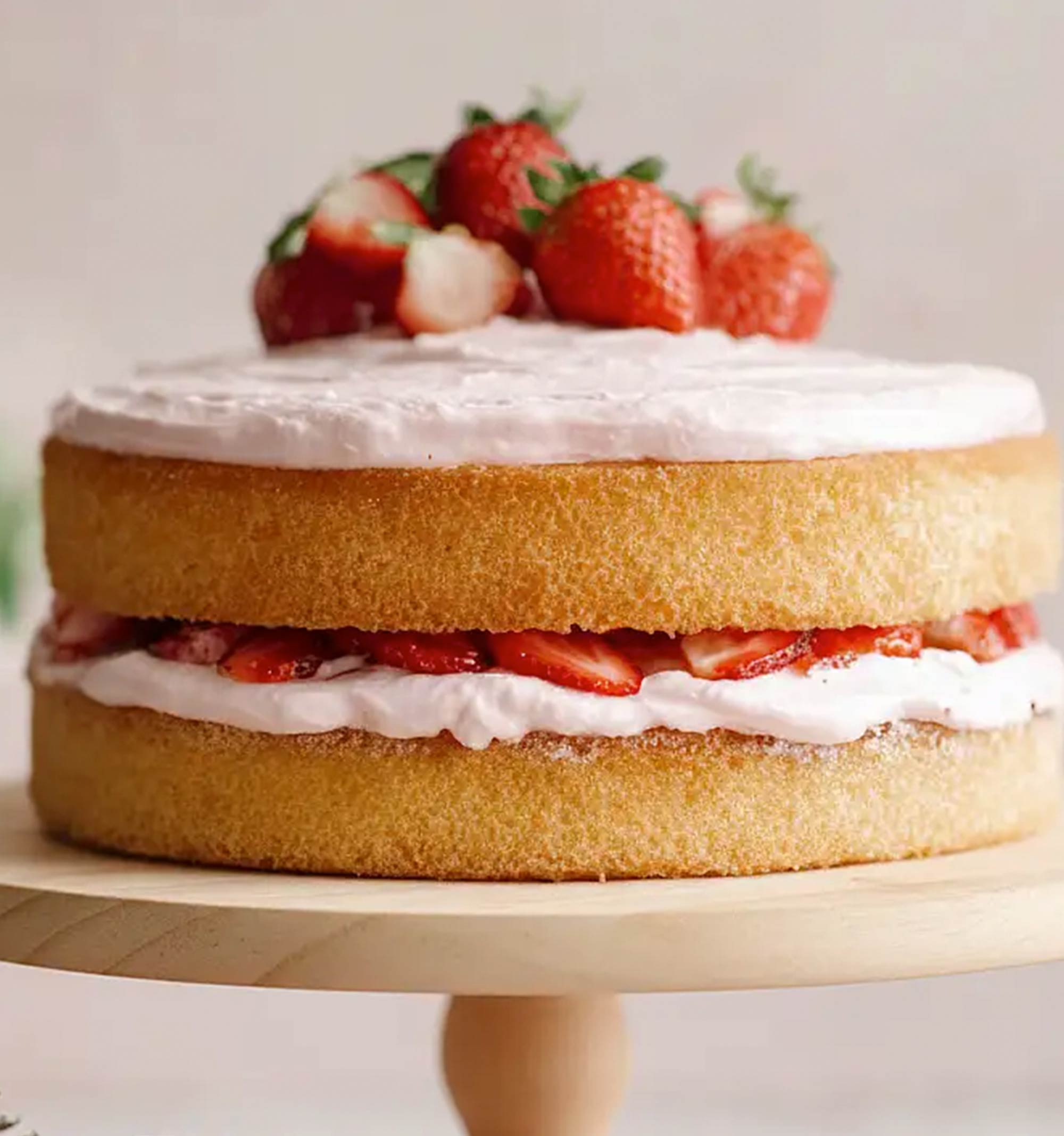 A two-layer cake sits on a wooden stand. A layer of white frosting and sliced fresh strawberries is visible between the cake layers. The top is covered with white frosting and whole fresh strawberries.