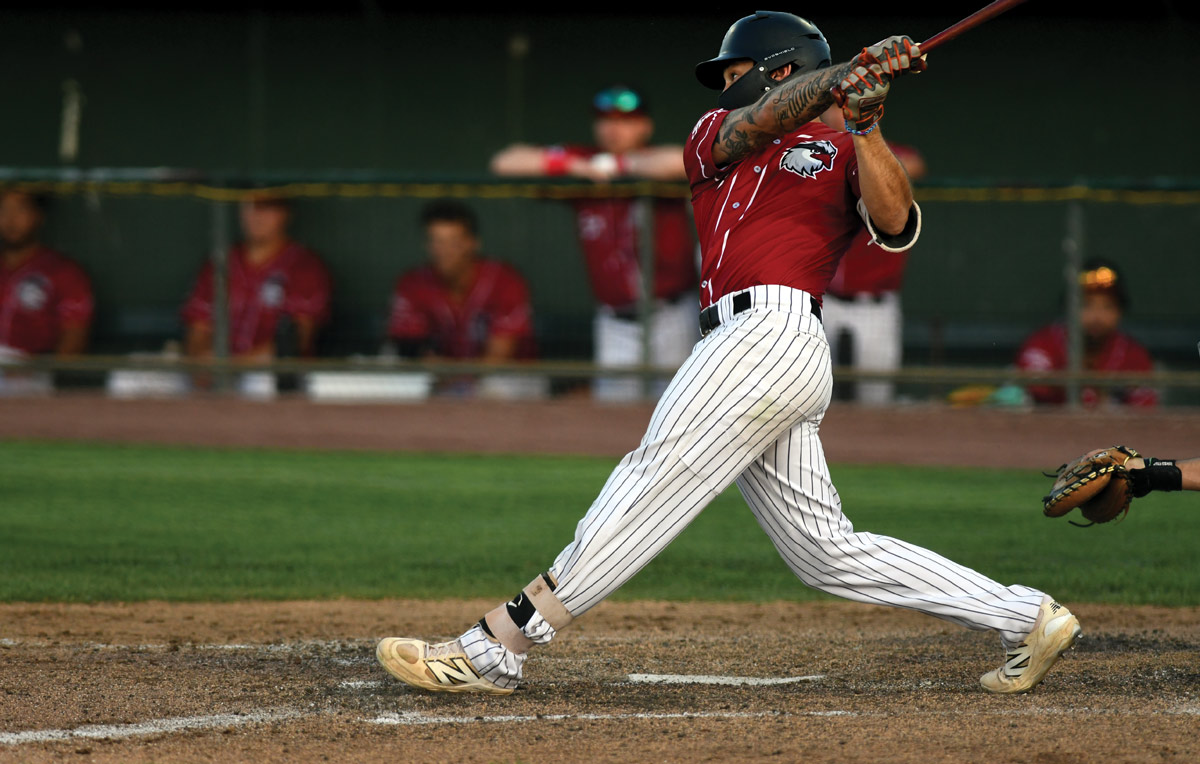 A baseball player in a red jersey and pinstriped pants swinging a bat during a game, with teammates visible behind him.