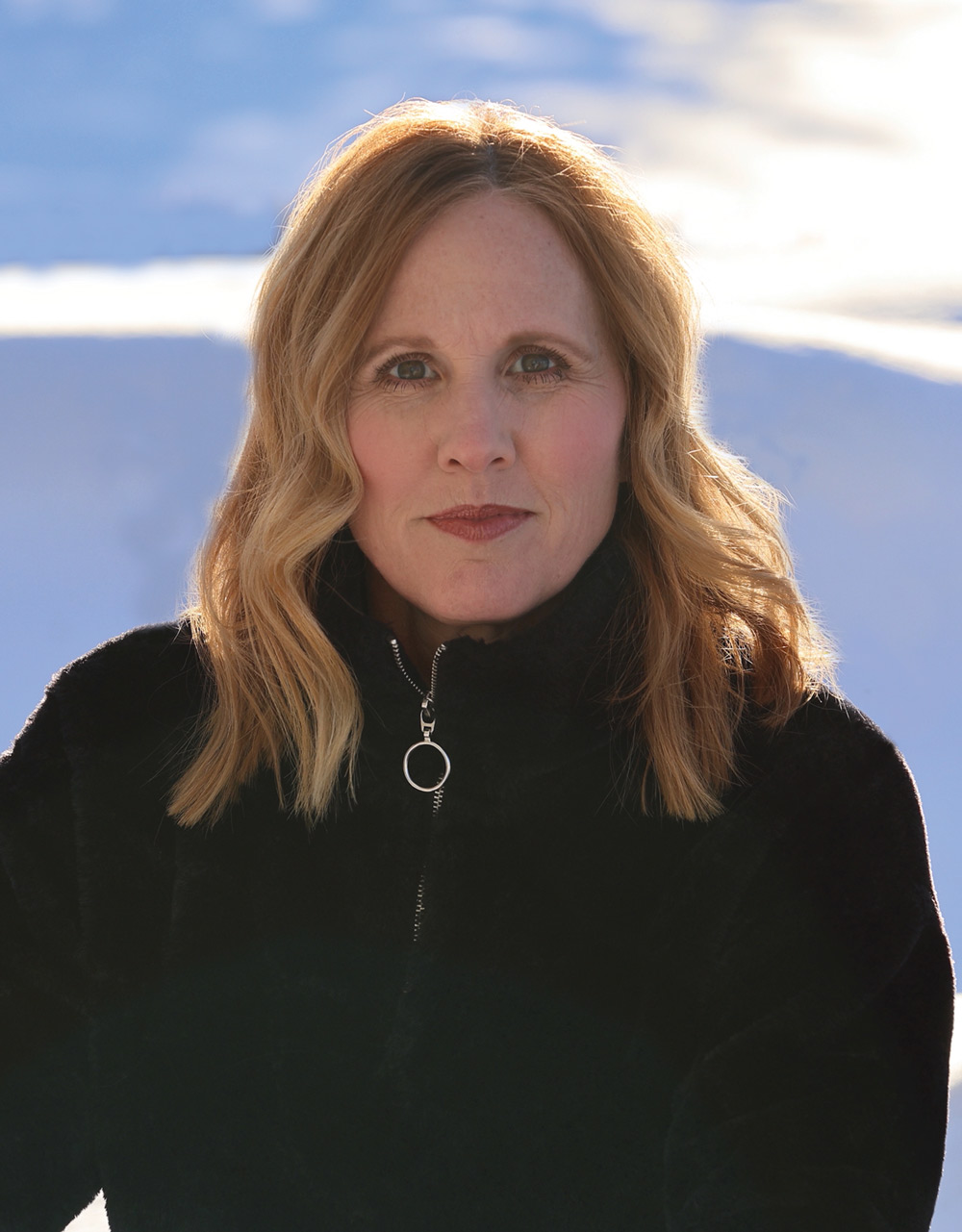 Portrait of a woman with shoulder-length blonde hair, wearing a black jacket, posing in bright outdoor lighting.