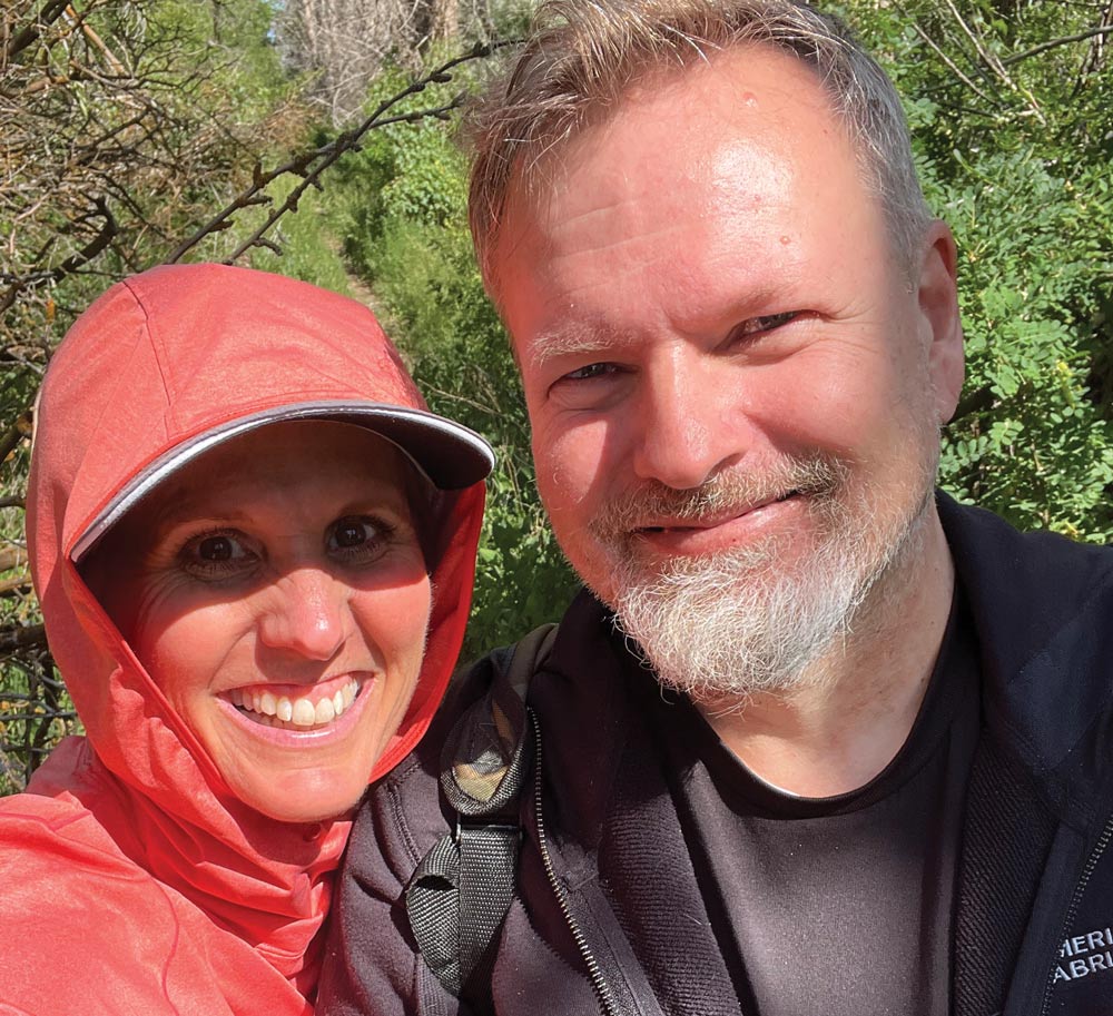 A smiling woman wearing a red hooded jacket and a man with a gray beard pose together for a selfie outdoors.