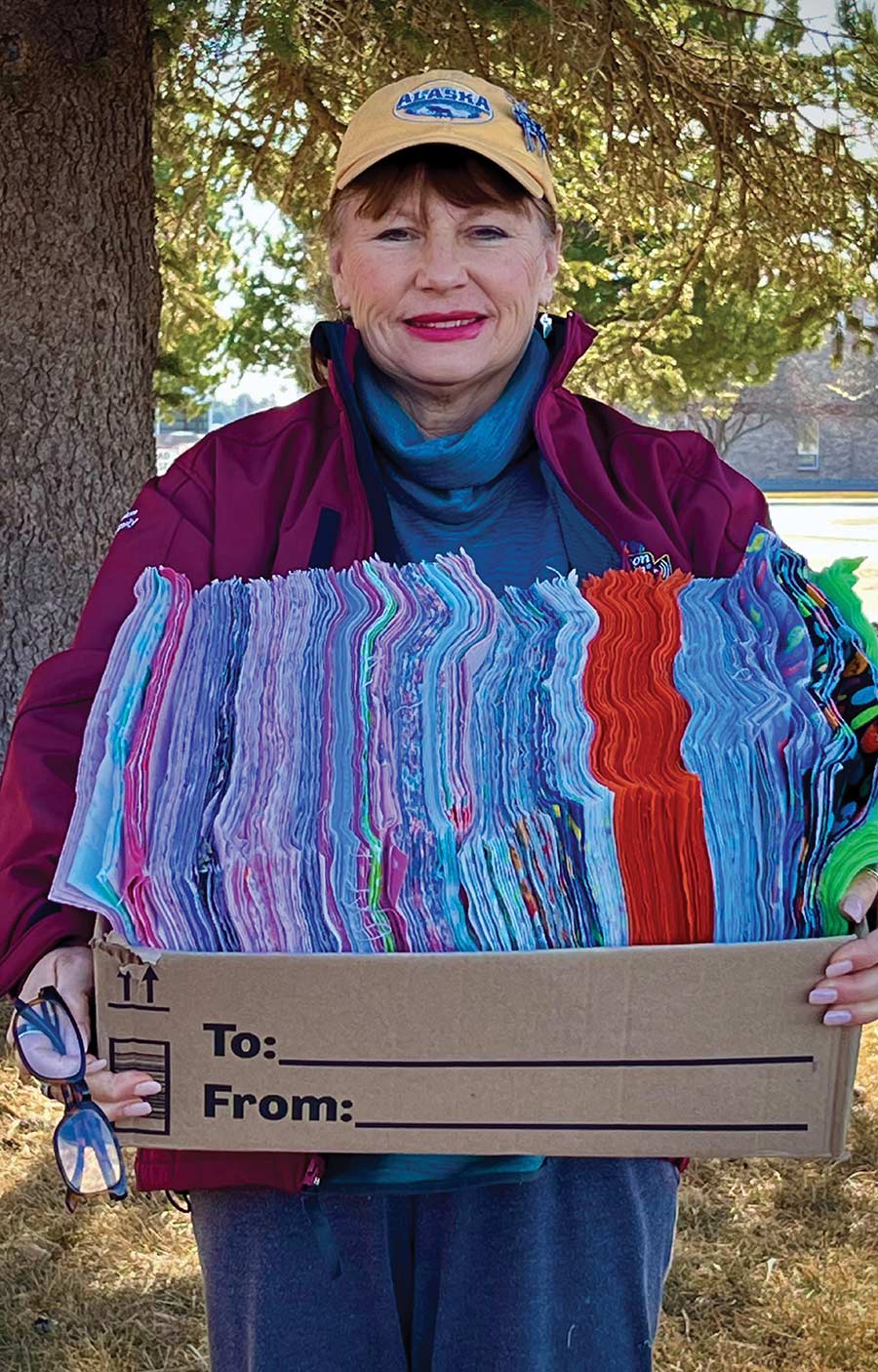 A woman smiles while holding a large cardboard box filled with stacks of colorful fabric.