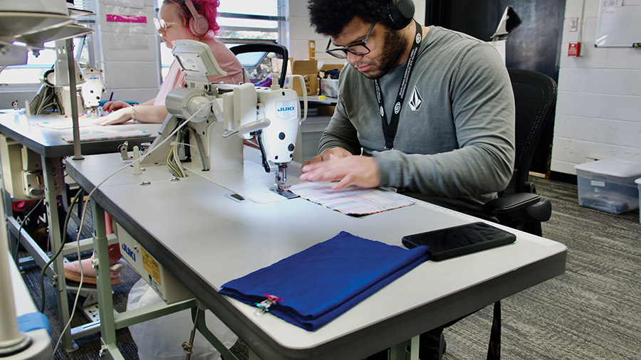 Two people working at industrial sewing machines stitching pieces of fabric in a workshop.