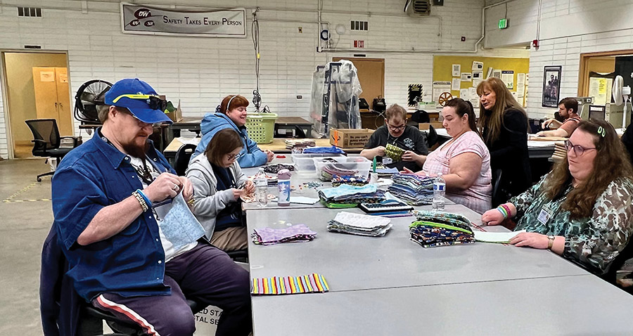 A group of people sitting around a long table, hand-sewing and organizing colorful fabric items