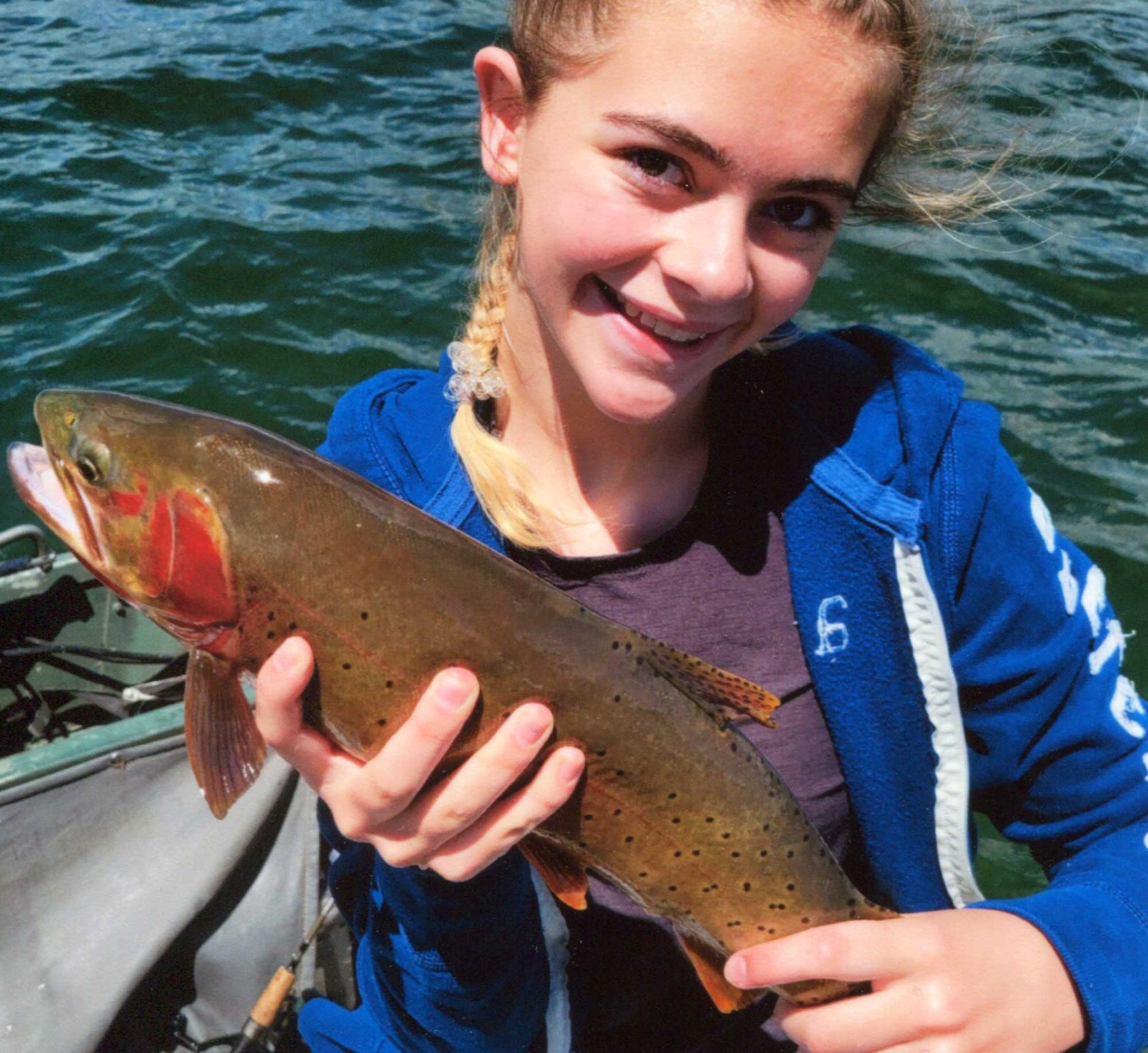 A smiling girl on a boat holds up a large trout she just caught.