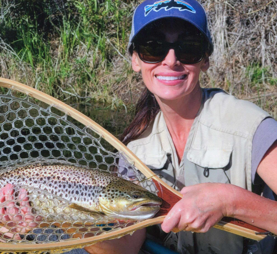 A woman in sunglasses and a hat holds a spotted trout in a fishing net.