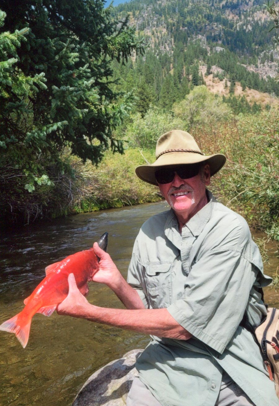 A man in a sun hat sits by a river holding a bright red salmon.