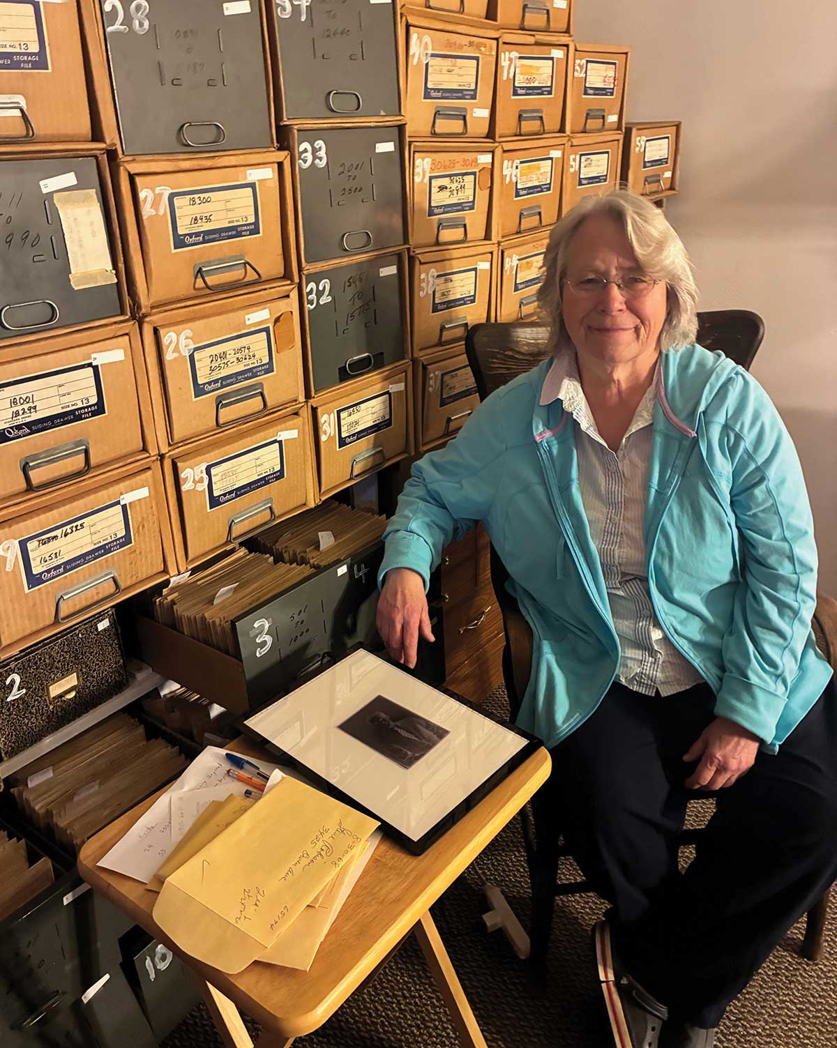 Reneta Adamson sits in a chair next to stacks of archived photo storage boxes.
