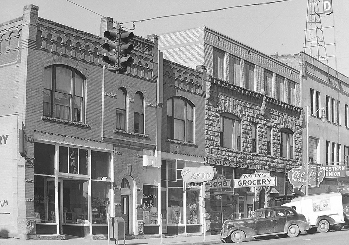 A black-and-white photo of storefronts including Wally's Grocery, The Spud Cafe, and Chesbro Music.