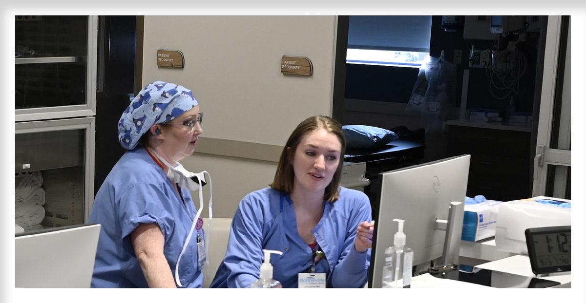 Amy Houston, wearing a patterned scrub cap, and Natalie Evans work together at a computer desk. Signs reading "Patient Recovery" are visible on the wall behind them.