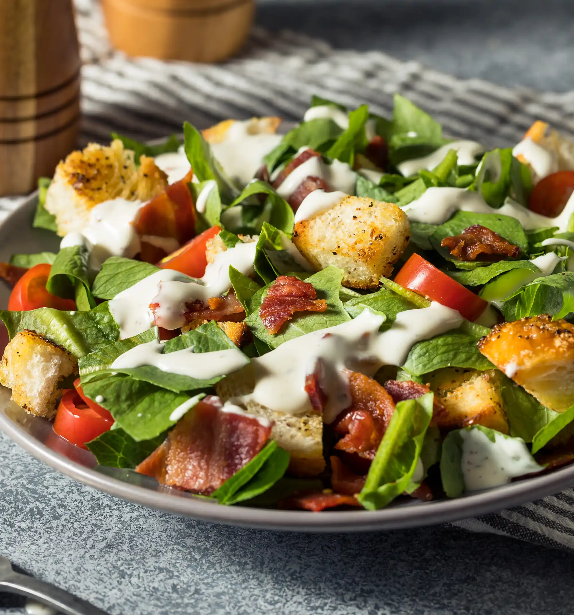 A chopped BLT salad in a grey bowl, consisting of green lettuce leaves, sliced tomatoes, pieces of bacon, and large toasted croutons, all drizzled with a creamy white dressing.