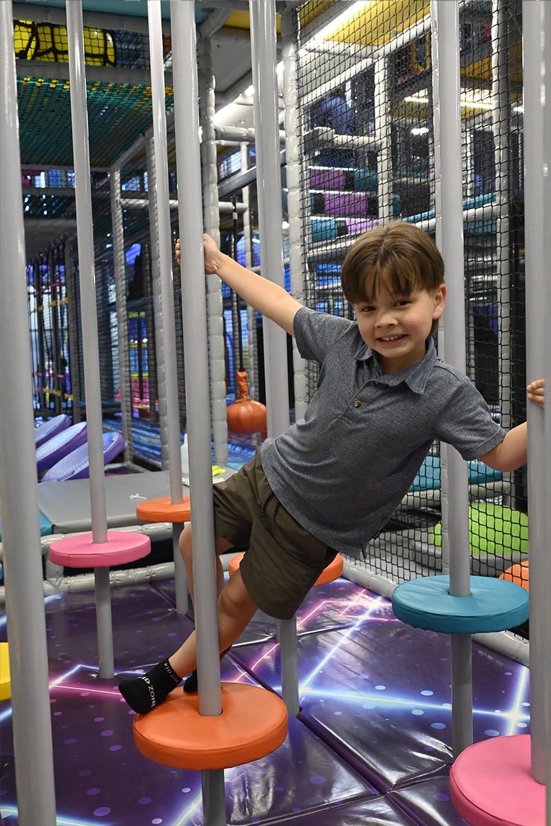 A smiling young boy navigates an indoor play structure, holding onto vertical metal poles and stepping across colorful circular pads.