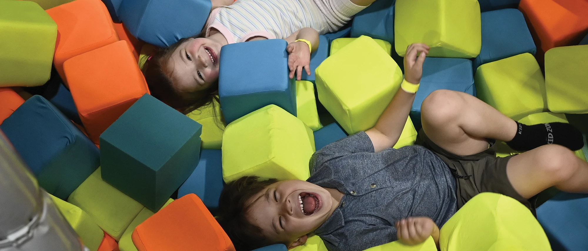 Two laughing children laying down and playing inside a pit filled with large, colorful foam blocks.