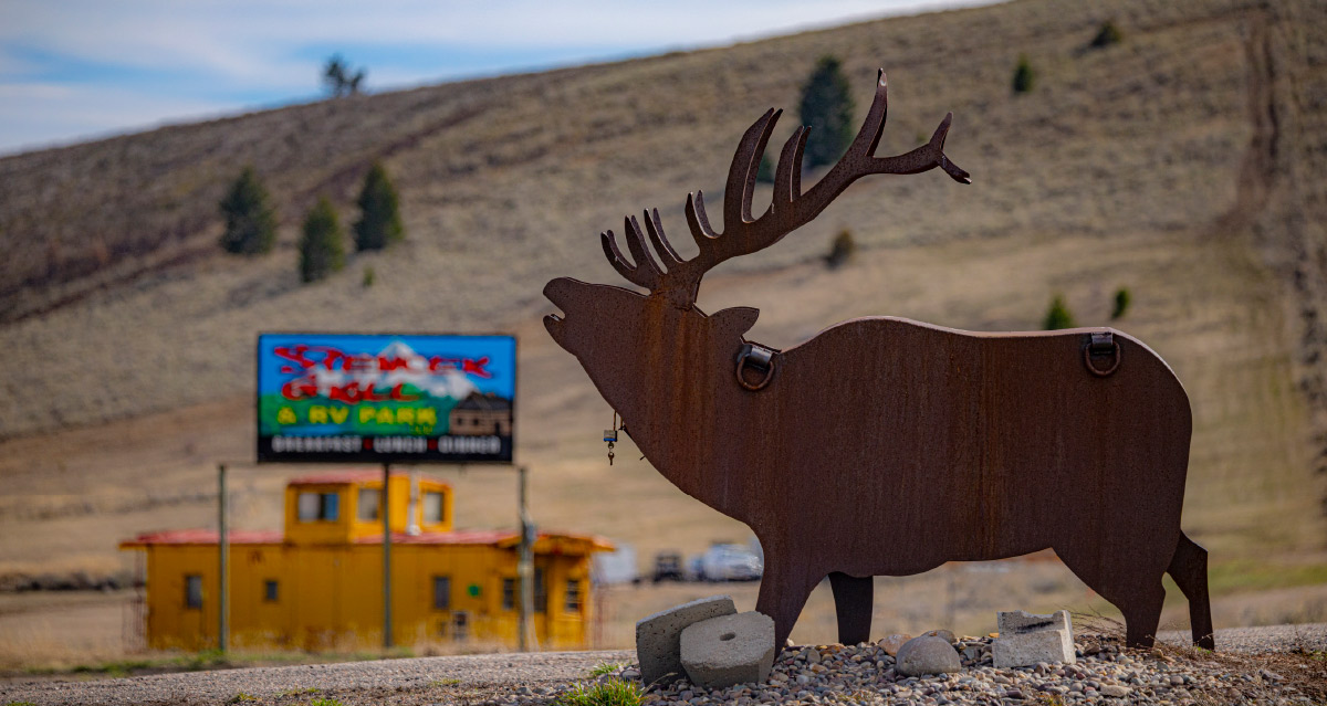 A rusty metal silhouette cutout of an elk standing near a roadside sign for an RV park, with a yellow train caboose in the background.