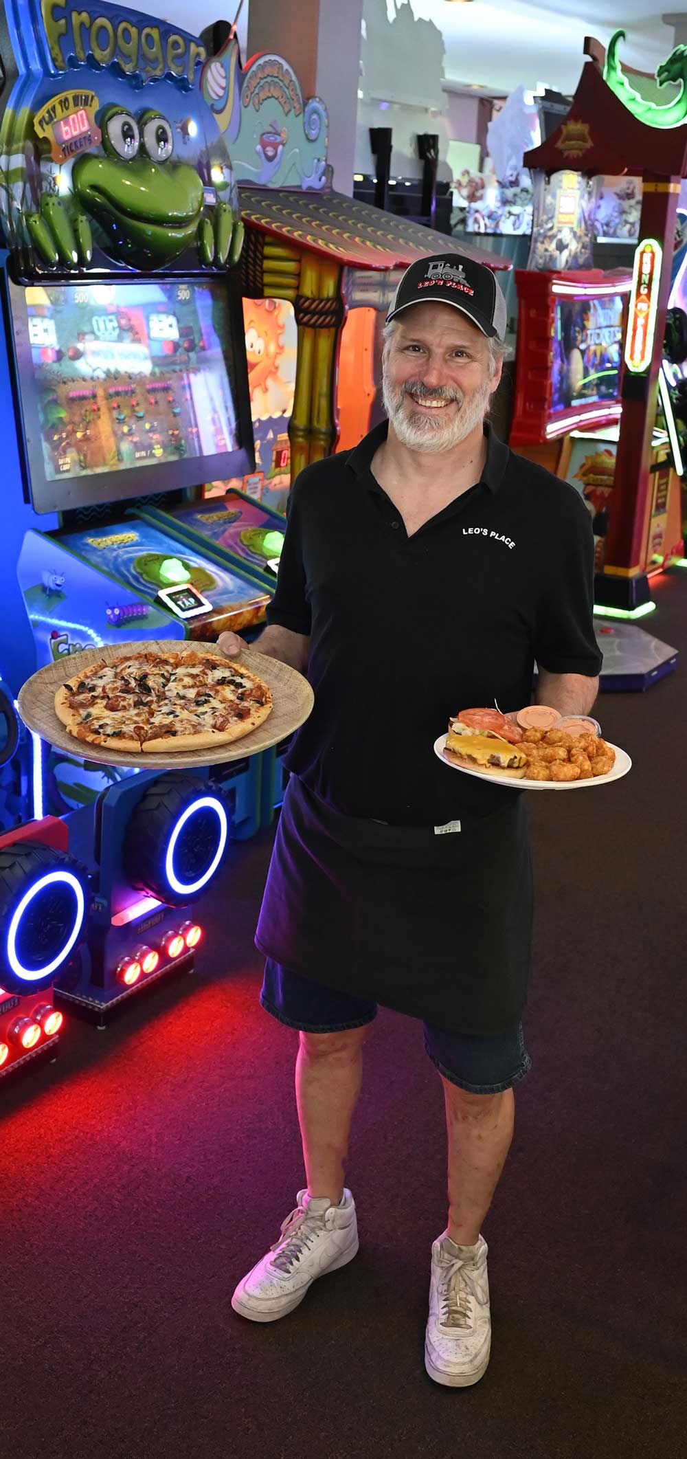A smiling man in a black polo shirt and cap holds a fresh pizza and a plate with a cheeseburger and tater tots inside a brightly lit arcade.